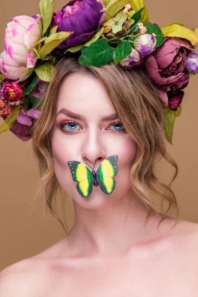 Нежная молодая женщина, позирующая в цветочном венке с искусственной зеленой бабочкой, изолированная на охетном фоне. бабочка, прикрывающая рот молодой женщины — стоковое фото