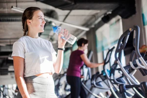 Smilende sportskvinne i hvit t0shirtholding flaske med vann i gymsalen . – stockfoto