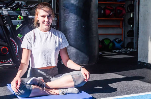 Дівчина в білій футболці і сірі легінси сидить на гімнастичному килимку в позі йоги в фітнес-клубі, посміхається і дивиться на камеру — стокове фото