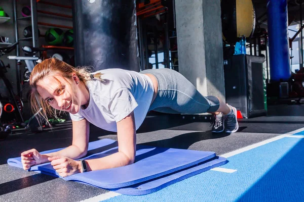 A girl in a white T-shirt and gray leggings sits doing the bar on a gymnastic mat in a fitness club. Sunlight and hard shadows in a fitness club — 스톡 사진