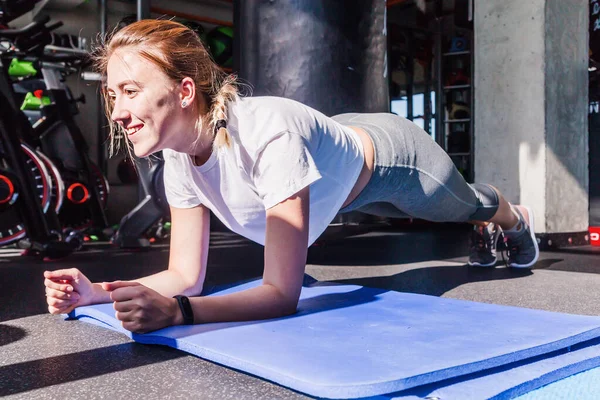 Дівчина в білій футболці і сірі легінси сидить, роблячи бар на гімнастичному килимку у фітнес-клубі. Сонячне світло і тверді тіні у фітнес-клубі — стокове фото