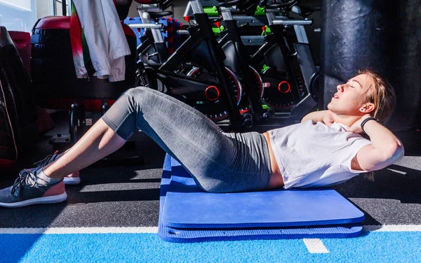 A girl in a white T-shirt and gray leggings sits doing an exercise on the abdominal muscles on a gymnastic mat in a fitness club. — 스톡 사진