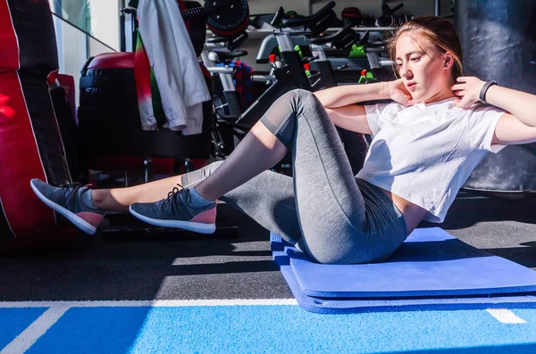 Дівчина в білій футболці і сірі легінси сидить, роблячи вправи на м'язах живота на гімнастичному килимку в фітнес-клубі . — стокове фото