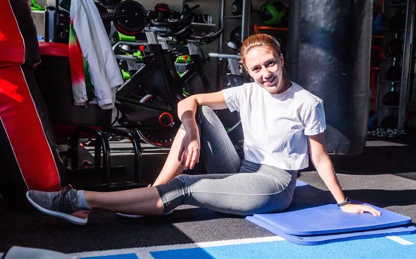 Дівчина в білій футболці і сірі легінси сидить на гімнастичному килимку у фітнес-клубі, посміхається і дивиться на камеру . — стокове фото