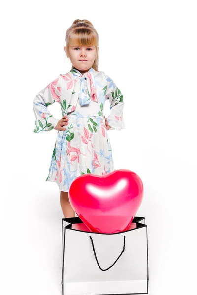 Una niña de moda en un vestido blanco con un estampado floral se encuentra junto a un gran paquete con un globo en forma de corazón en el interior —  Fotos de Stock
