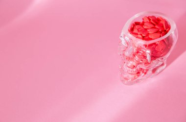İnsan kafatası şeklinde bir cam bardak kırmızı kalplerle doludur ve pembe arka planda durur.