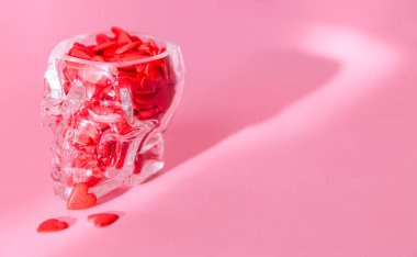 İnsan kafatası şeklinde bir cam bardak kırmızı kalplerle doludur ve pembe arka planda durur.