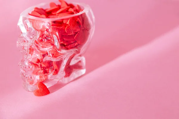 Egy emberi koponya alakú üvegpohár tele van vörös szívvel, és rózsaszín alapon áll. — Stock Fotó