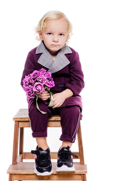 Petit garçon à la mode dans un élégant costume bordeaux et avec un bouquet de fleurs à la main sur un fond blanc. Le garçon est triste, contrarié, en colère — Photo