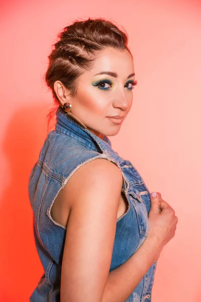 Beautiful brunette girl with make-up in blue tones, pigtails and a sleeveless jeans vest posing against a red background — Stok fotoğraf
