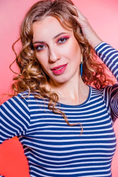 Menina morena bonita feliz com maquiagem vermelha, cabelo encaracolado e uma camisola listras azuis posando contra um fundo vermelho . — Fotografia de Stock