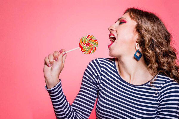 Happy beautiful brunette girl with red makeup, curly hair and a blue striped sweater, posing against a red background with a colored lollipop in her hands. Girl pretending to sing