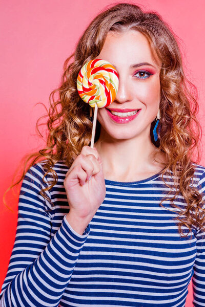 Happy beautiful brunette girl with red makeup, curly hair and a blue stripes sweater posing against a red background. One eye is covered with candy