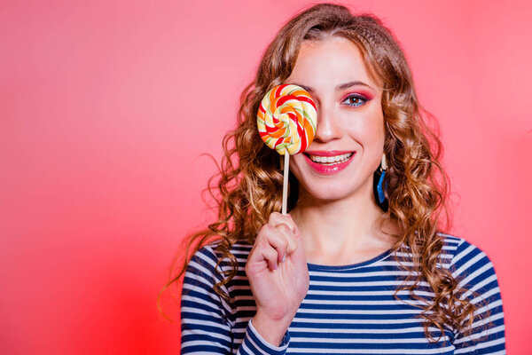 Happy beautiful brunette girl with red makeup, curly hair and a blue stripes sweater posing against a red background. One eye is covered with candy