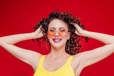 Kıvırcık saçlı, sıradan sarı tişörtlü, kolları ve boynu arkasında esneyen, kırmızı arka planda mutlu bir şekilde gülümseyen genç bir kız.