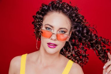 Kıvırcık saçlı, sarı tişörtlü, kırmızı arka planda poz veren güzel, mutlu esmer kız.