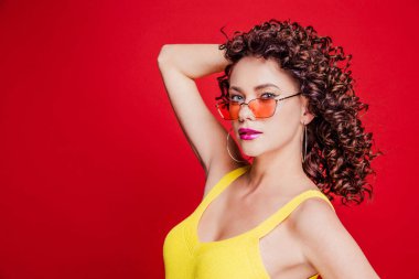 Kıvırcık saçlı, sarı tişörtlü, kırmızı arka planda poz veren güzel, mutlu esmer kız.