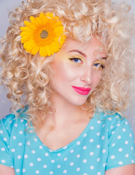Beautiful blonde girl with voluminous curly hairstyle, in a blue polka dot blouse and with a flower of a sunflower in her hair on a gray background — Stock Photo, Image