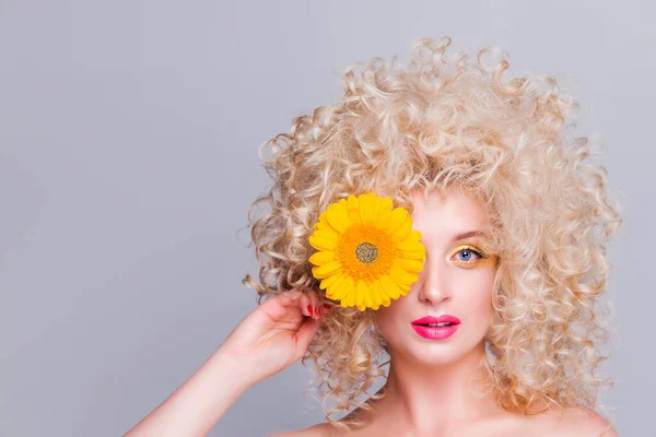 Beautiful fashionable blonde girl with voluminous curly hairstyle, bare shoulders and a sunflower in her hands on a grey background. woman looking at camera and covering eye with a flower — Stock Photo, Image