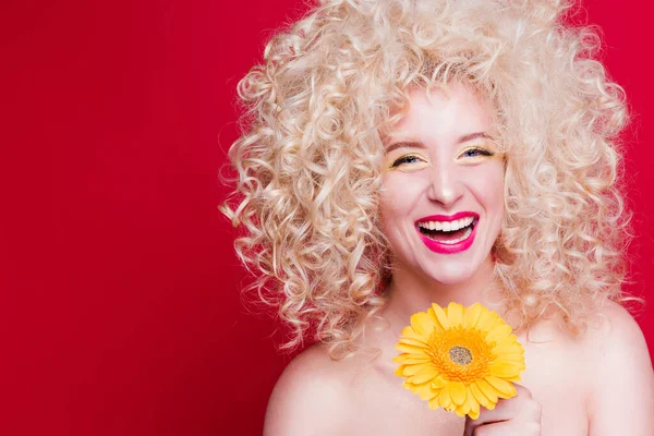 Beautiful fashionable blonde girl in retro style with voluminous curly hairstyle, bare shoulders and a sunflower in her hands on a red background. Happy girl laughing — Stock Photo, Image