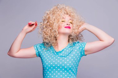 Gri bir arka planda mavi benekli bluzu olan, eski moda kıvırcık saçlı güzel sarışın kız, saç stilinden hoşlanıyor ve kameraya bakıyor.
