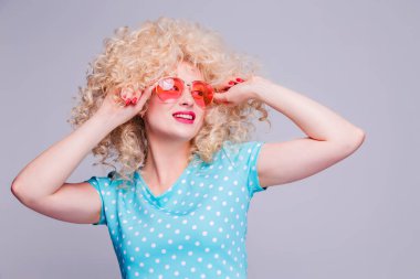 Gri bir arka planda mavi benekli bluz ve pembe gözlüklü, kıvırcık saçlı, eski tarz sarışın bir kız.