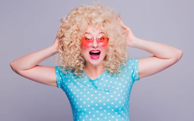 Gri bir arka planda, şehvetli kıvırcık saçlı, mavi benekli bluzlu ve pembe gözlüklü, geçmişe dönük güzel sarışın kız neşe ve haz yaşıyor.