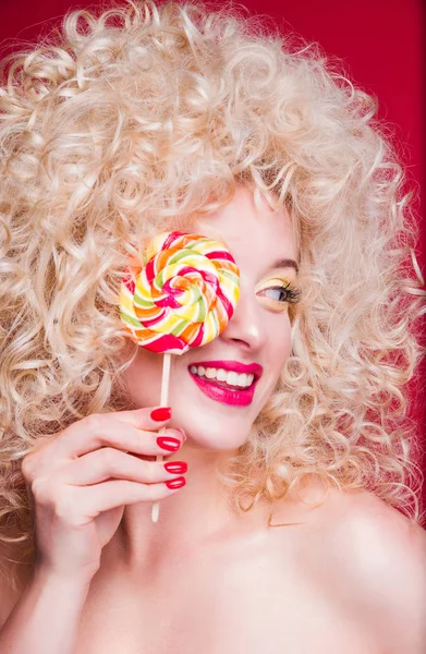 Beautiful fashionable blonde girl in retro style with voluminous curly hairstyle, open shoulders on a red background smiles, laughs and closes one eye with colored candy — Stock Photo, Image