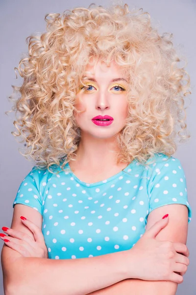 Mooi modieus blond meisje in retro stijl met volumineus krullend kapsel, in een blauwe polka dot blouse op een grijze achtergrond kijkt naar de camera — Stockfoto