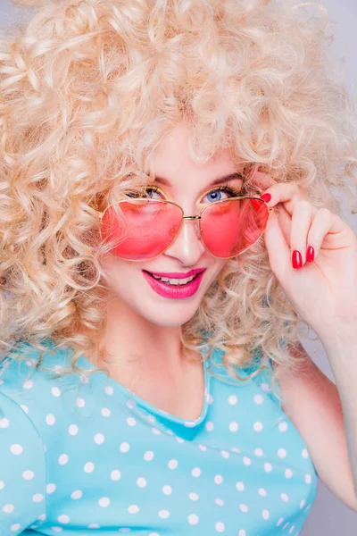 Beautiful retro-style blonde girl with voluminous curly hairstyle, in a blue polka-dot blouse and pink glasses on a gray background