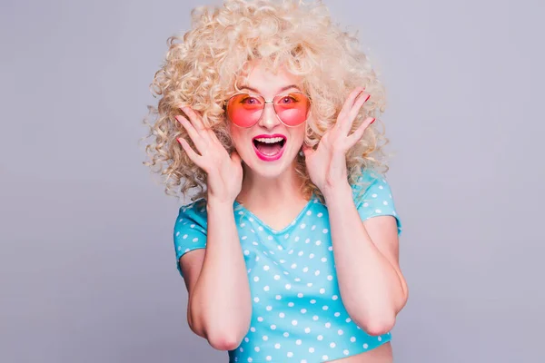 Beautiful blonde girl in retro style with voluminous curly hairstyle in a blue polka-dot blouse and pink glasses on a gray background experiences joy and delight — Stock Photo, Image