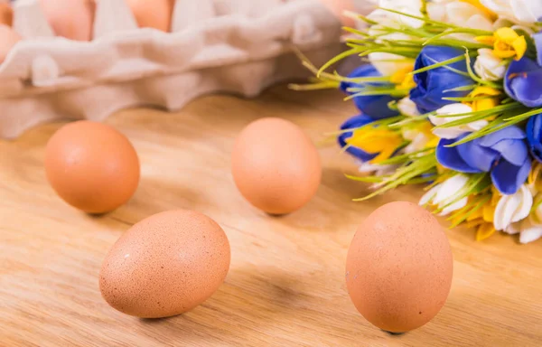 Brown eggs in packaging and without packaging, a bouquet of yellow and blue flowers lie on a wooden surface. Top view