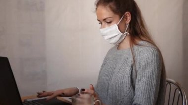 Beyaz koruyucu maskeli bir kız masada oturuyor, dizüstü bilgisayarla çalışıyor ve telefonla konuşuyor. Serbest çalışma, uzaktan çalışma ve karantina kavramı. video