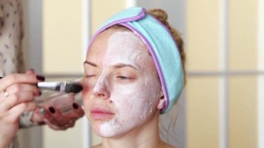 Bir makyaj sanatçısı makyaj yapmadan önce bir kızın yüzüne nemlendirici maske takar. Prosedür evde gerçekleşiyor. Video