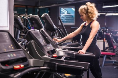 Spor salonunda koşu bandında siyah spor kıyafetli bir kız. Kız ekranda bir düğmeye basıyor. Yatay fotoğraf