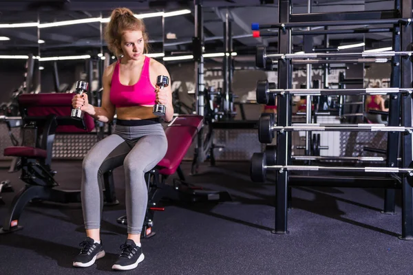 Girl posing in the gym with dumbbells in hands, sitting on a bench. Horizontal photo