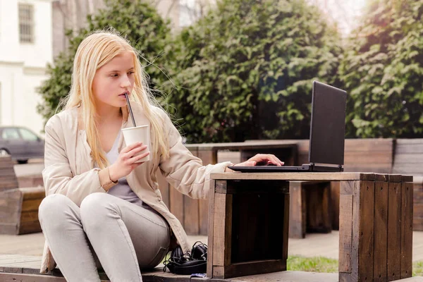 一个小女孩坐在咖啡店的街上，喝咖啡，看笔记本电脑 — 图库照片