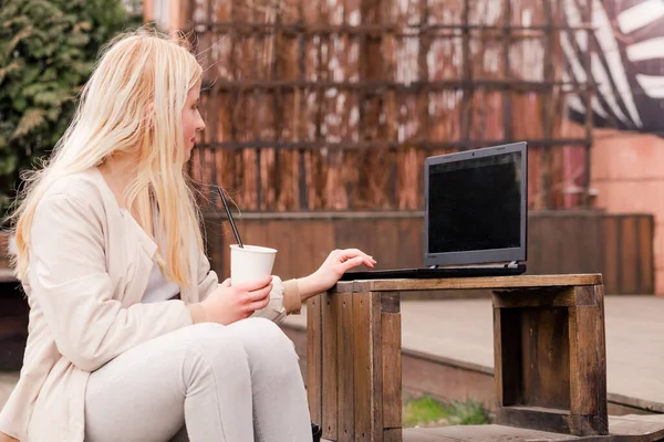 Ein junges Mädchen sitzt in einem Café auf der Straße, trinkt Kaffee und blickt auf einen Laptop — Stockfoto
