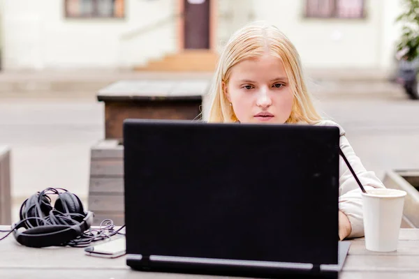 Ein junges Mädchen sitzt draußen in einem Café mit einem Laptop. Das Mädchen schaut ernsthaft auf den Laptop-Bildschirm. — Stockfoto