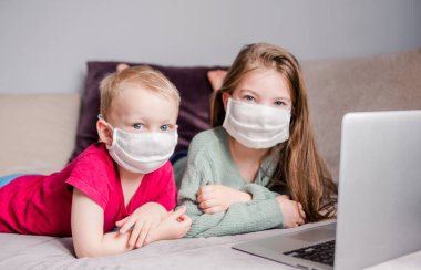İki çocuk, beyaz tıbbi maskeler takmış bir kanepede yatıyorlar ve çocukların yanında bir dizüstü bilgisayar olan kameraya bakıyorlar. Karantina, kendini izole etme ve salgın hastalık kavramı. Yatay fotoğraf