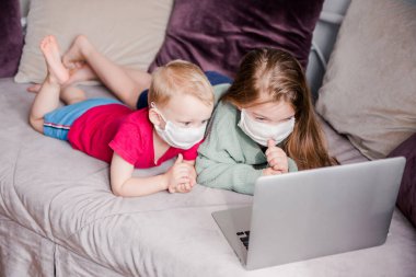 İki çocuk evlerindeki bir kanepede beyaz sağlık maskeleriyle yatıp dizüstü bilgisayarlarında video izliyorlar. Karantina, kendini izole etme ve salgın hastalık kavramı. Üst görünüm