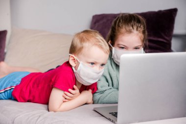 İki kız ve erkek kardeş dizüstü bilgisayar ve evde eğitim kullanarak tıbbi maskelerle kanepede yatıyorlar. Evde kalıyorlar. Yatay fotoğraf