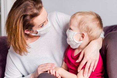Beyaz tıbbi maskeli anne ve oğul birbirlerine bakıyorlar. - Evde. Yatay fotoğraf