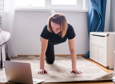 Siyah eşofmanlı güzel bir kadın evde egzersiz yapıyor ve dizüstü bilgisayarında video izliyor. Yatay fotoğraf