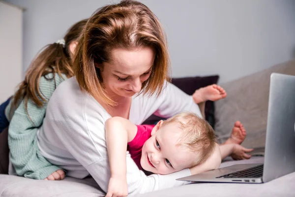 Young women works at home with laptop along with two children.Children want to communicate with mother, make noise and interfere with work.Self-isolation during coronovirus pandemic. Horizontal photo