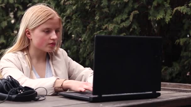 一个年轻姑娘坐在街上的咖啡店里 旁边放着笔记本电脑 耳机和电话 — 图库视频影像