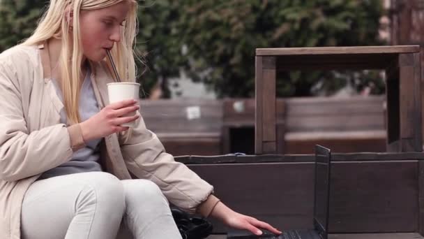一个年轻的女孩坐在咖啡店的街上 喝咖啡 看笔记本电脑 — 图库视频影像
