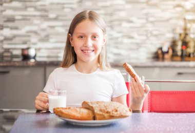 Mutfakta okul çağındaki bir kız oturuyor. Bir elinde bir bardak süt, diğer elinde bir parça taze ev yapımı ekmek var. Yatay fotoğraf