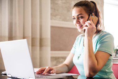 Genç bayan bir serbest çalışan uzaktan kumandayla evden çalışıyor. Masada ev kıyafetleriyle oturan ve telefonda konuşan bir kadın. Yatay fotoğraf