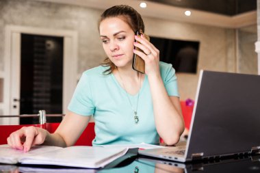 Genç bayan bir serbest çalışan uzaktan kumandayla evden çalışıyor. Bir kadın masa başında ev elbiseleriyle oturuyor, belgelere bakıyor ve telefonda konuşuyor. Yatay fotoğraf